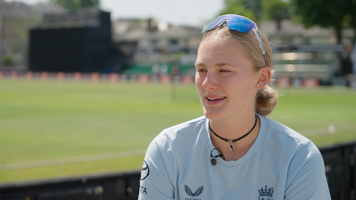 Tilly Corteen-Coleman, an England Women's cricketer, looks at a cricket bat whilst standing in a bright, sunny cricket ground.